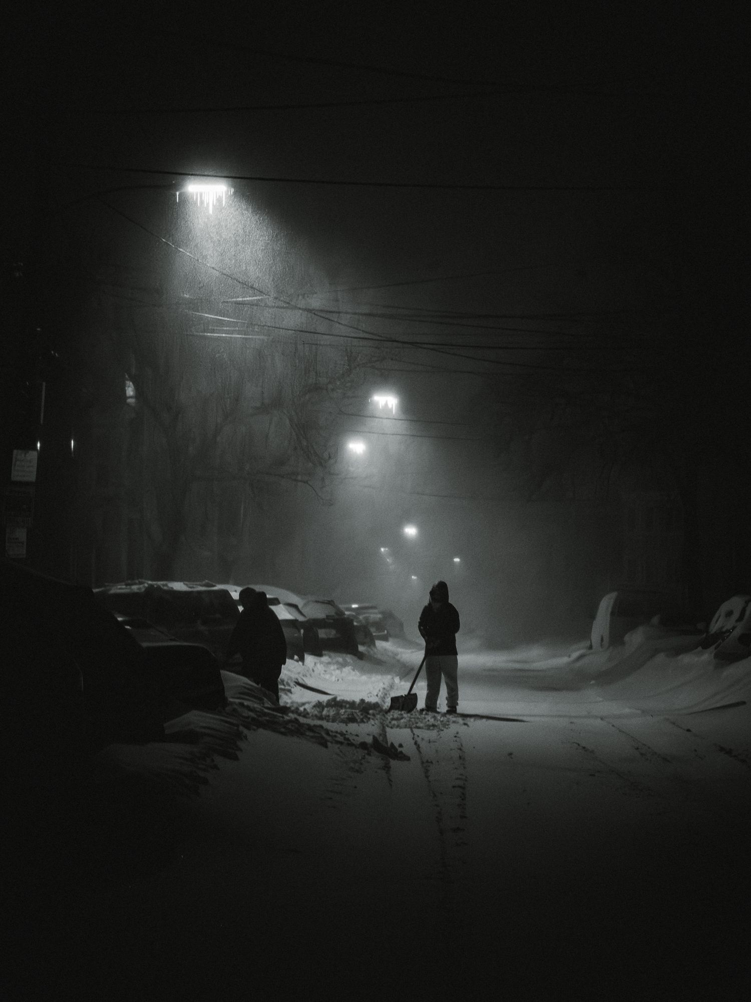Silhouettes of people and equipment are visible in the dark as snow falls under bright streetlights creating an eerie atmosphere.