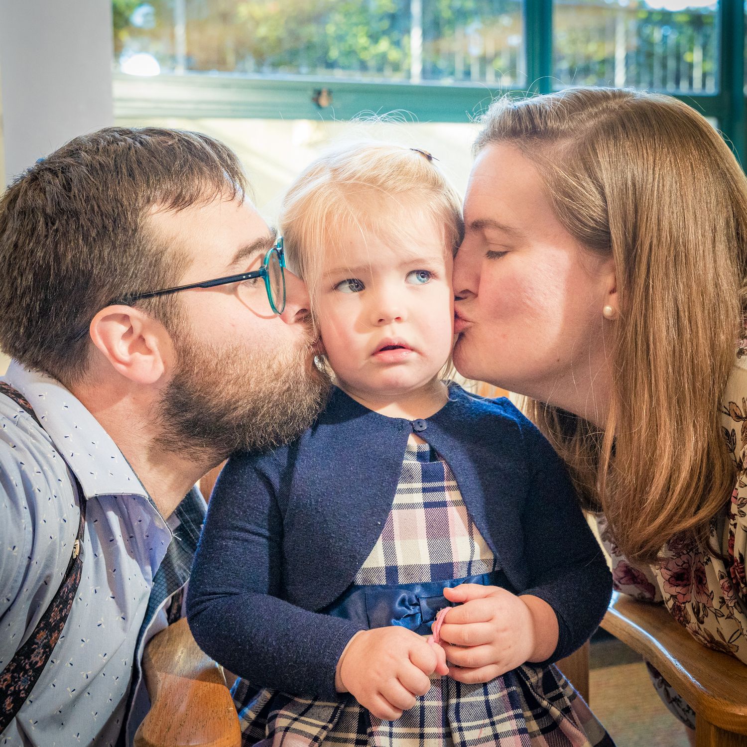 Parents giving kisses to a toddler in a navy dress and plaid pattern while sitting near a window.