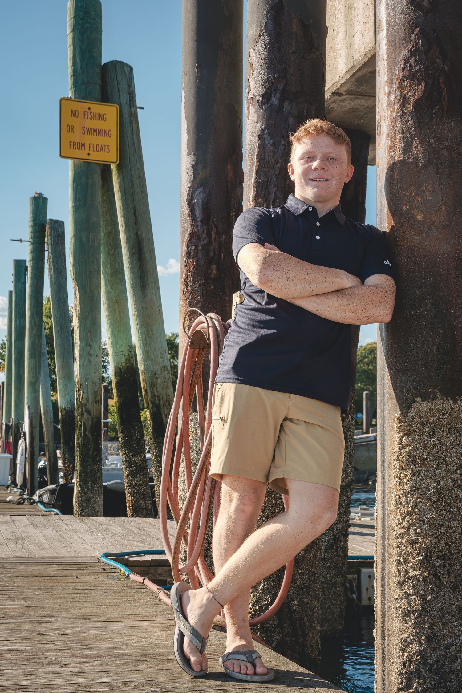 Young man with red hair leaning against pier post, holding rope coil, wearing navy polo and khaki shorts at marina.