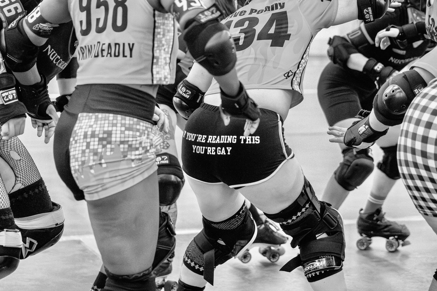 Black and white photo of roller derby skaters in action, wearing protective gear and numbered jerseys during a bout.