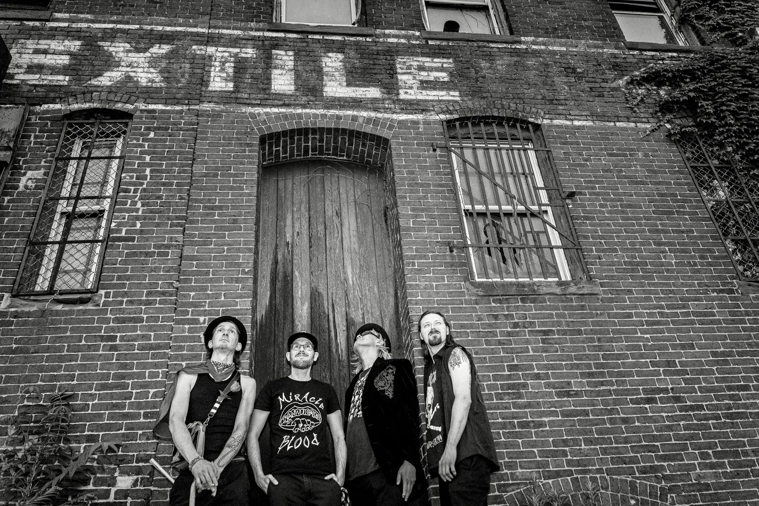 A group of musicians poses together outside a brick building wearing black clothing and matching outfits.