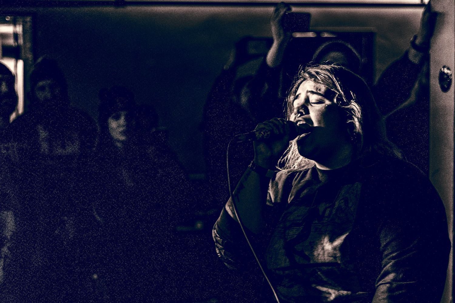 A dramatic portrait in dark moody lighting shows a performer passionately singing on stage in black and white.