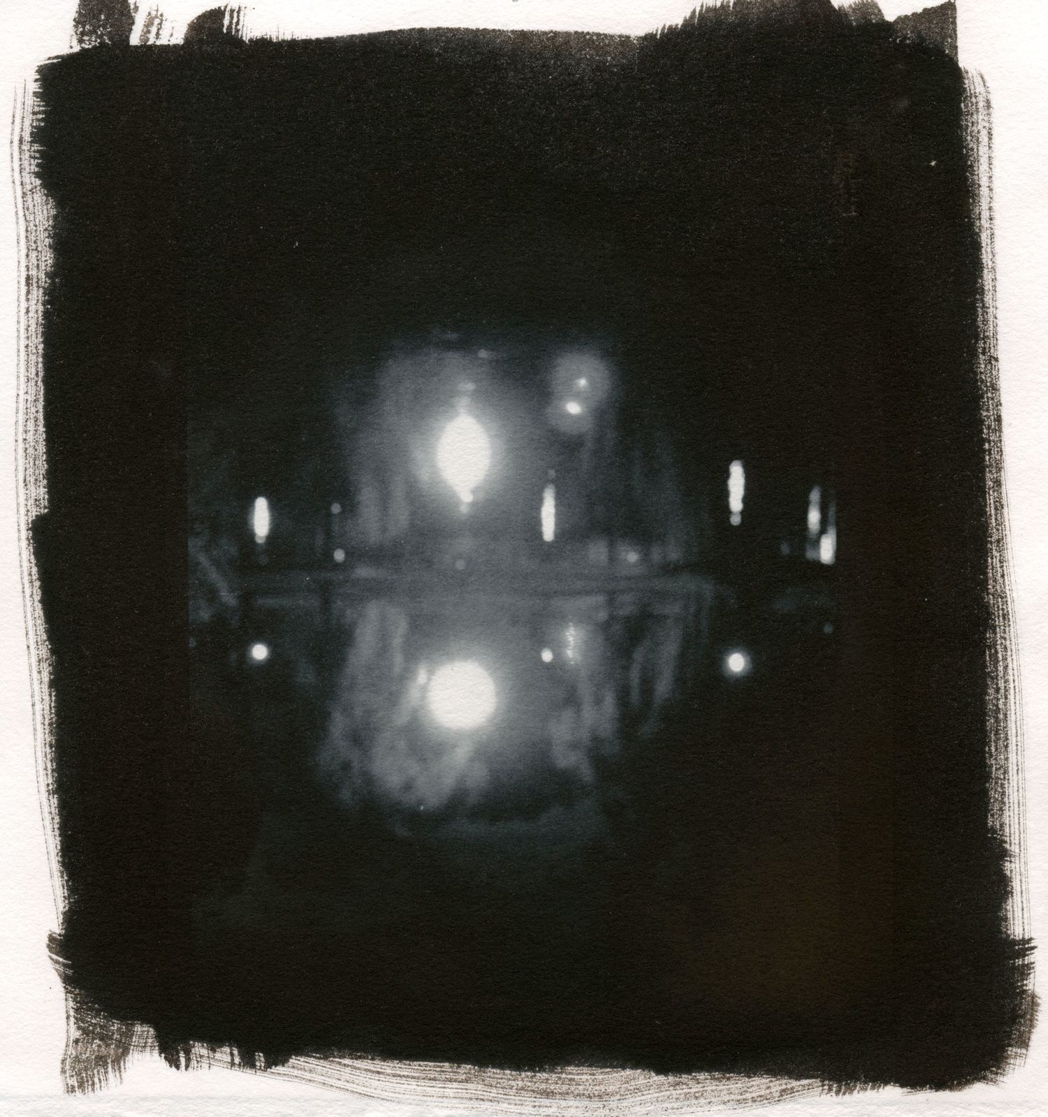 Moody black and white night scene with glowing street lights reflecting on water's surface, surrounded by dark borders.