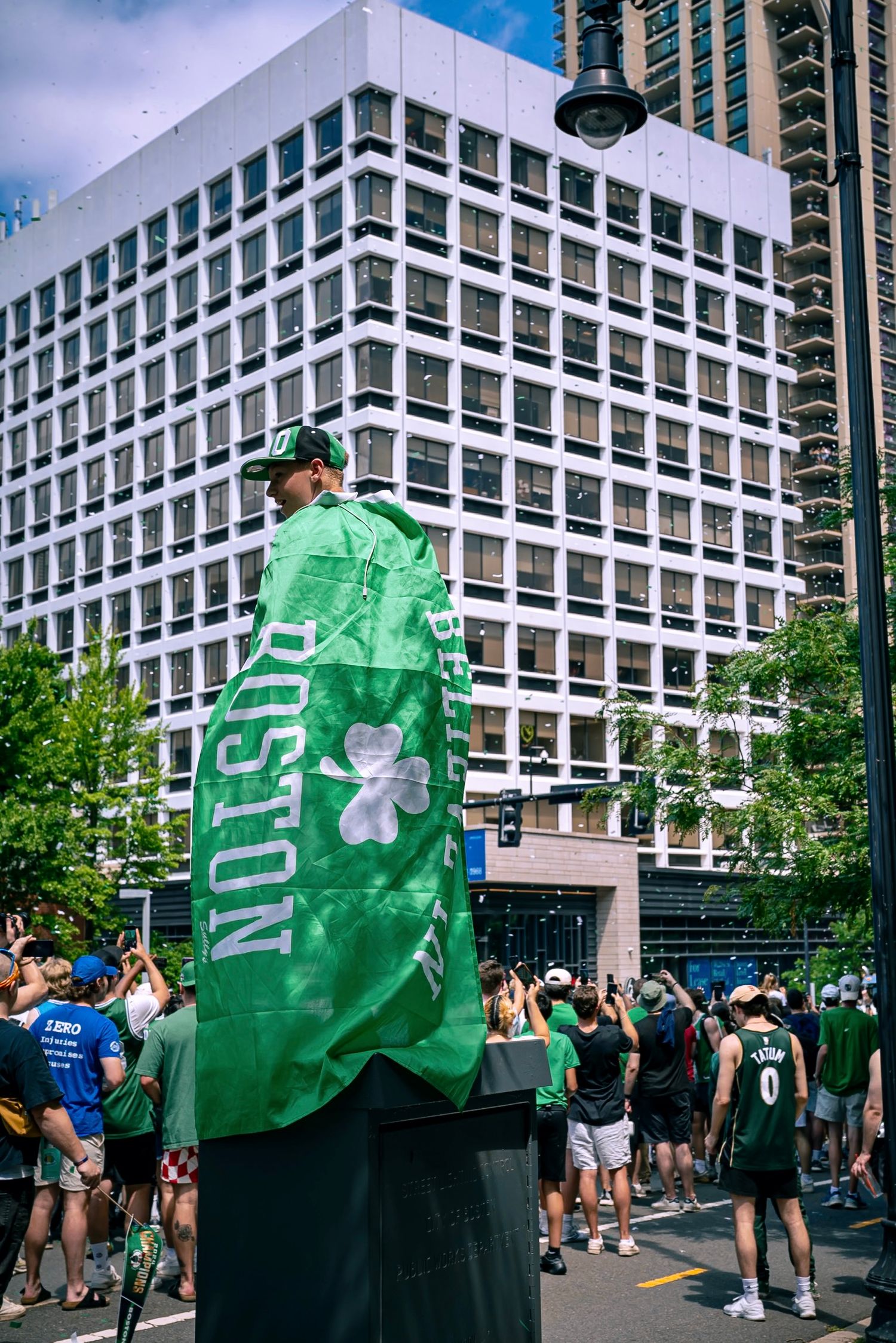 A person wearing a green Boston-themed flag stands in front of a modern office building during what appears to be a celebration.