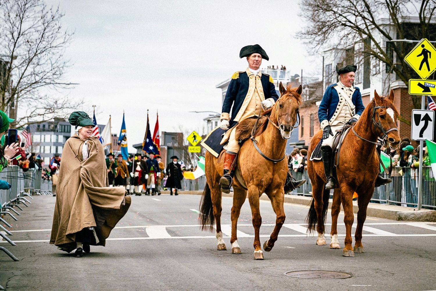 A group of riders on horseback in colonial period costumes parade along a city street during a historical reenactment event.