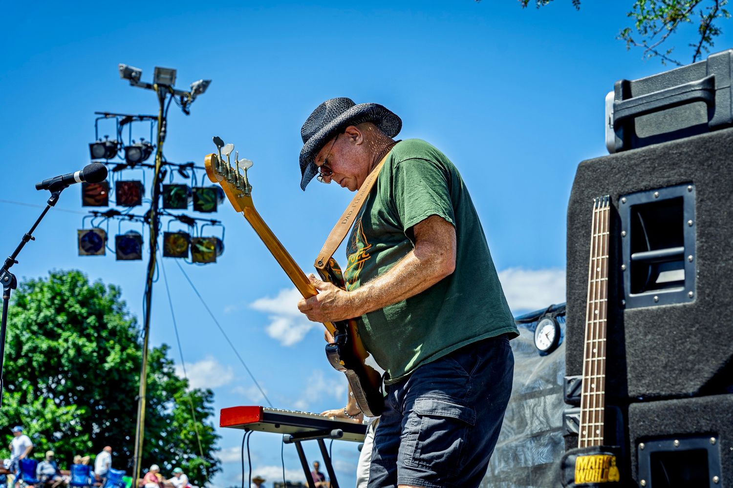 Electric guitarist performs on outdoor stage with sound equipment and speakers on sunny summer day.