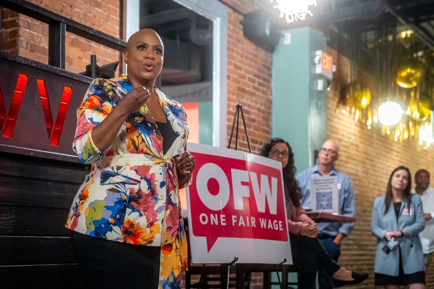 Individual in colorful dress speaks at One Fair Wage event with red logo and brick wall backdrop.