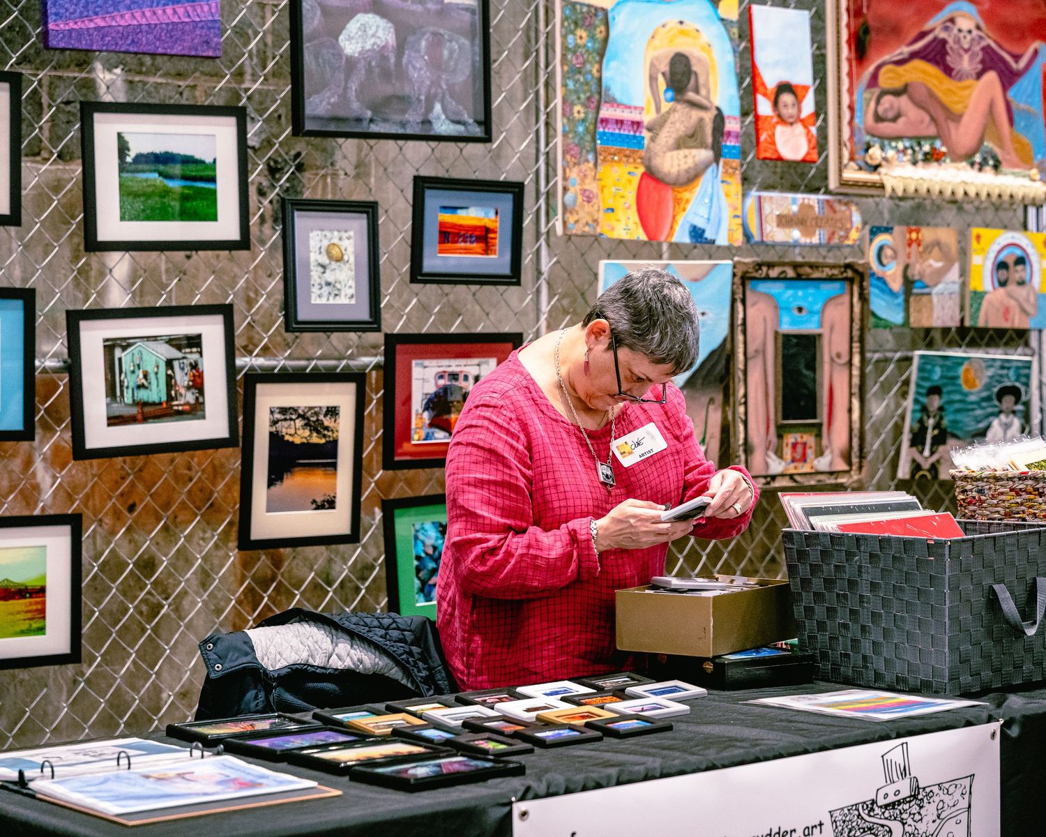 Art vendors display colorful artwork and framed pieces at an outdoor market booth with chain link fencing.