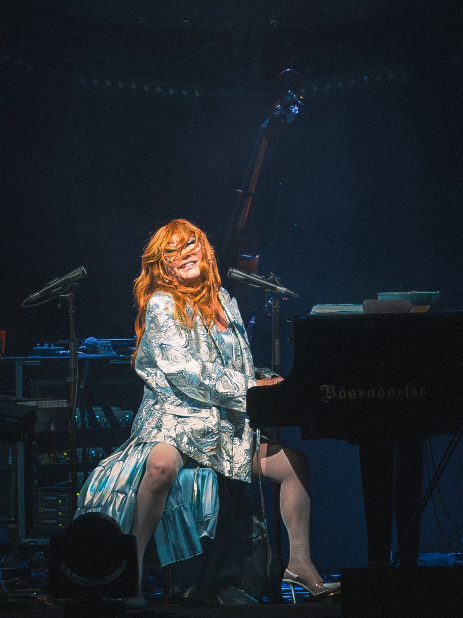 Red-haired woman in floral dress smiling while playing a Bösendorfer grand piano on a dimly lit concert stage.