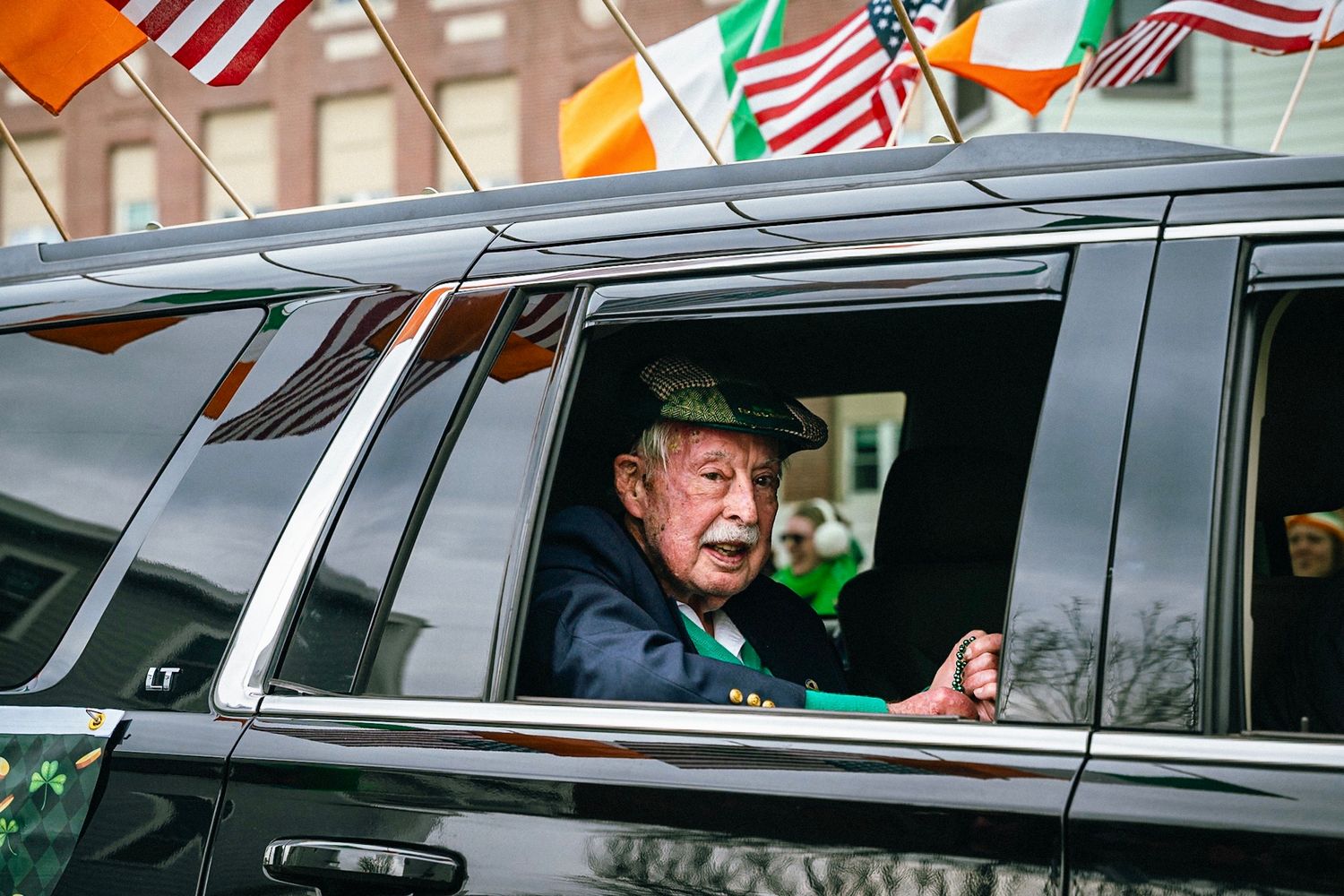 A line of black SUVs decorated with American and Irish flags drives in a parade procession through city streets.