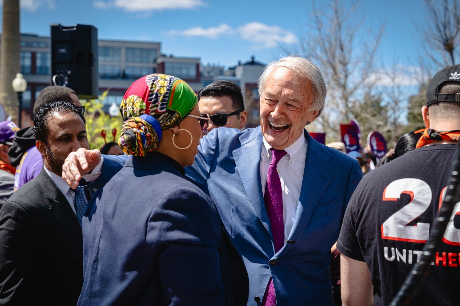 Politician in blue suit greeting diverse crowd at outdoor rally on sunny day.