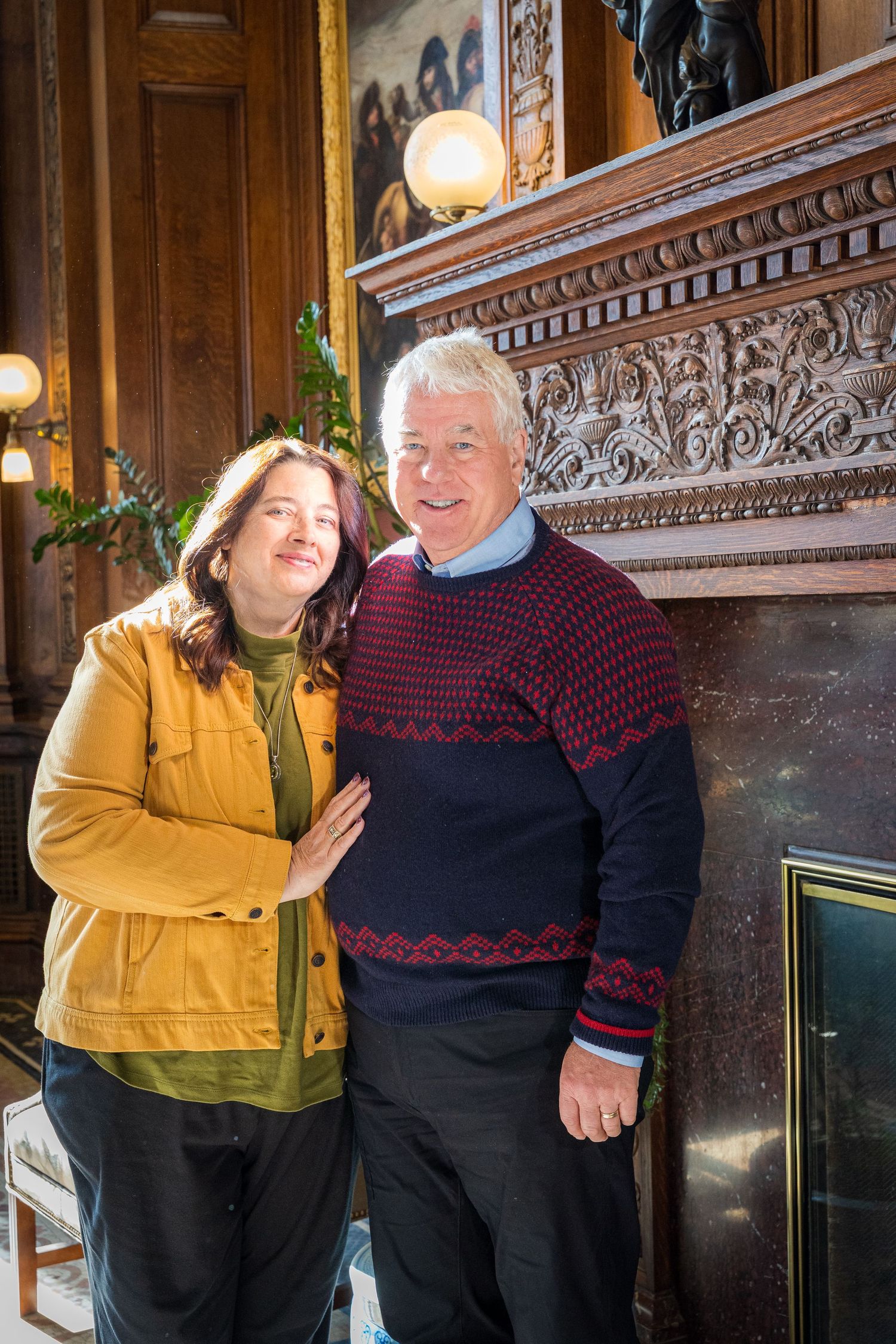 Two people smiling together beside an ornate wooden fireplace mantel in an elegant interior setting.