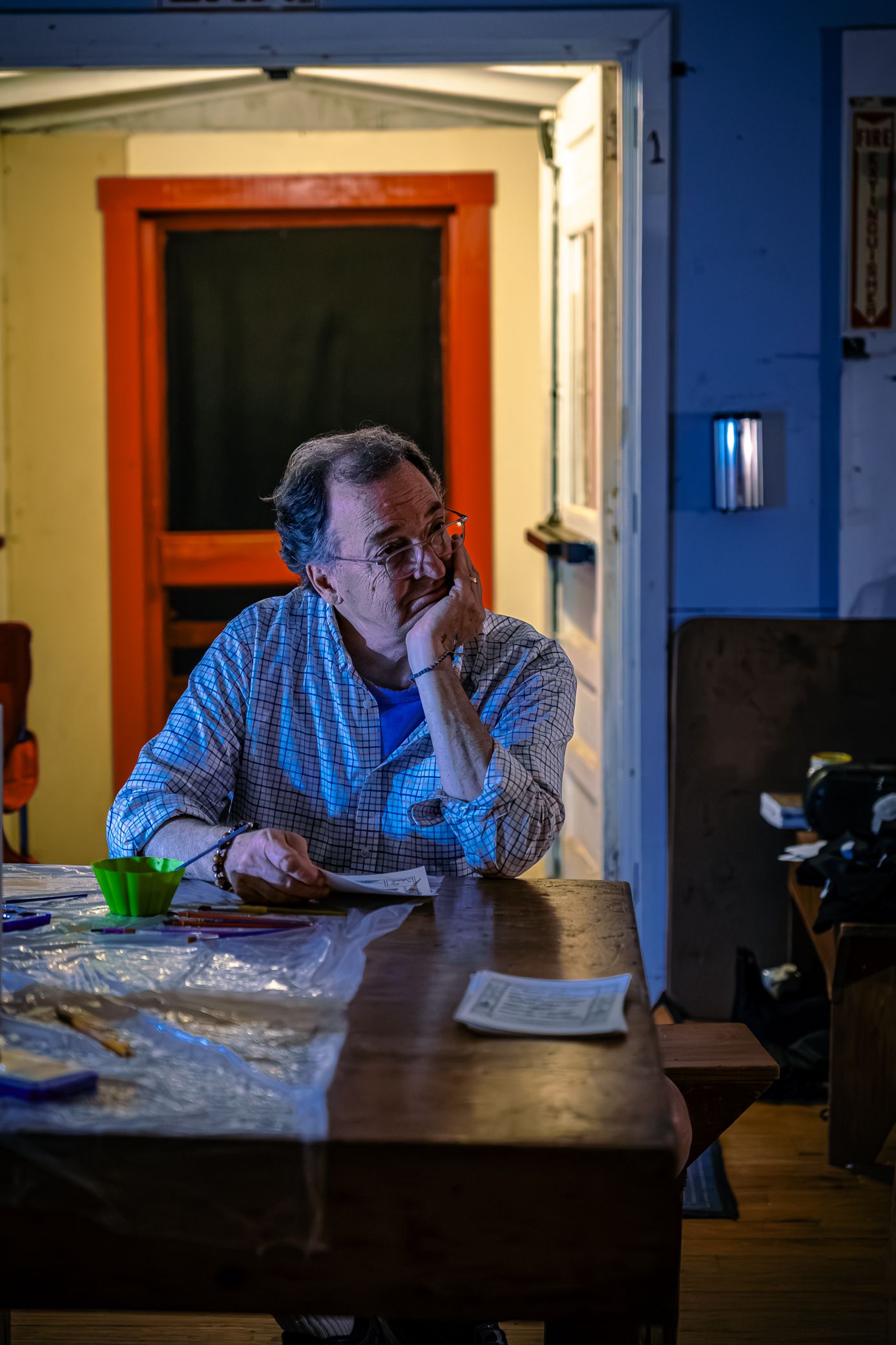 Person in plaid shirt sits at wooden table in evening light near doorway.