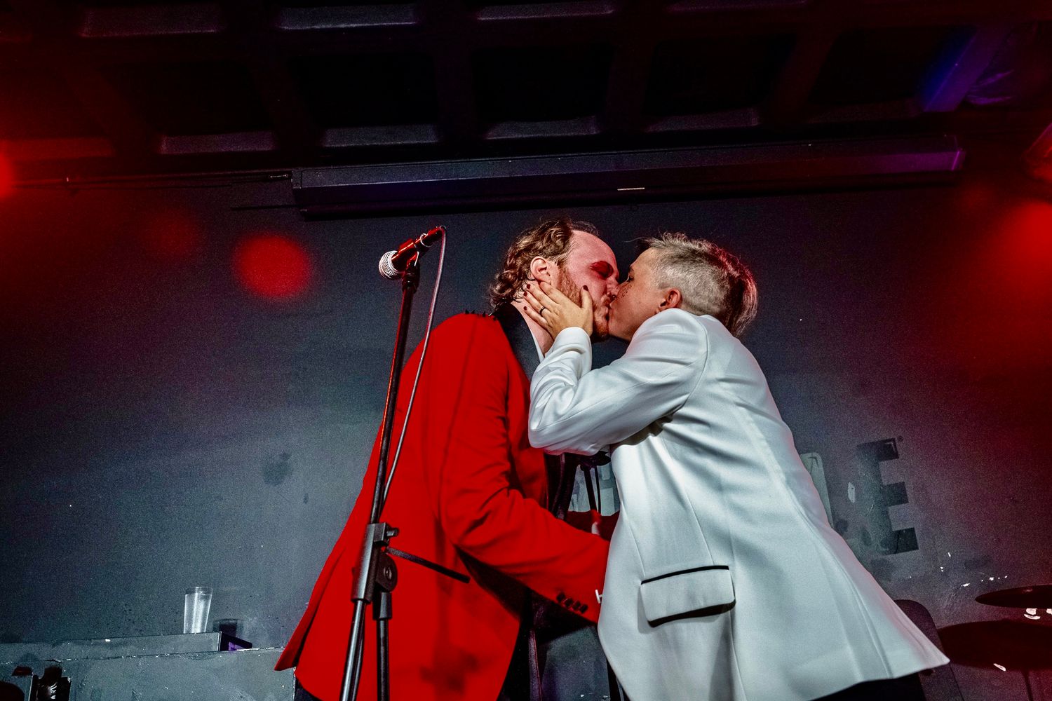A performer in a white jacket sings passionately on stage with dramatic red lighting in the background.