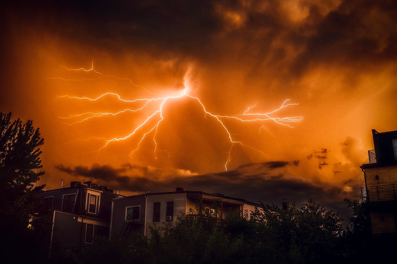 Dramatic lightning storm illuminates orange sky over suburban houses with dark storm clouds at night.