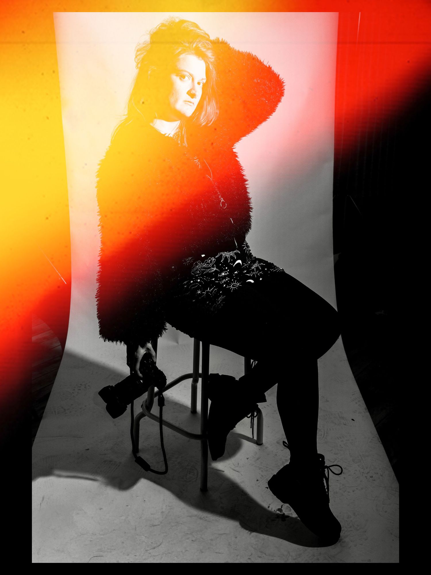 Woman in fur coat sits on stool with dramatic red and orange light effects in black and white studio photo.