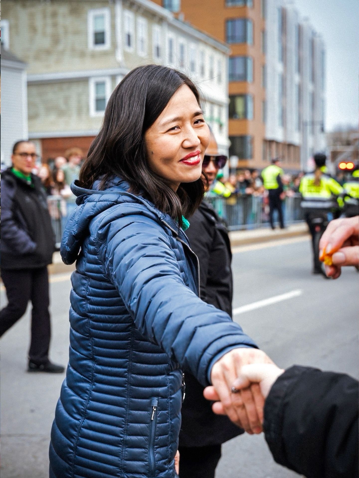 A person in a blue puffy jacket greets supporters at an outdoor event with crowd barriers and security visible.