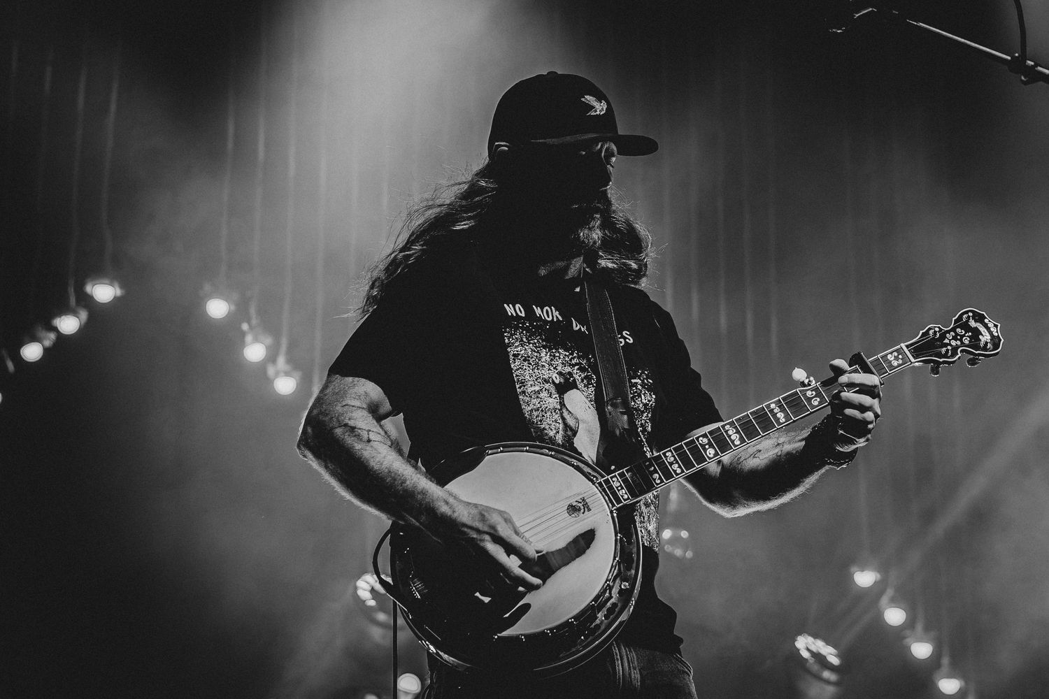 Black and white photo of a bearded musician playing banjo on stage under dramatic concert lighting.