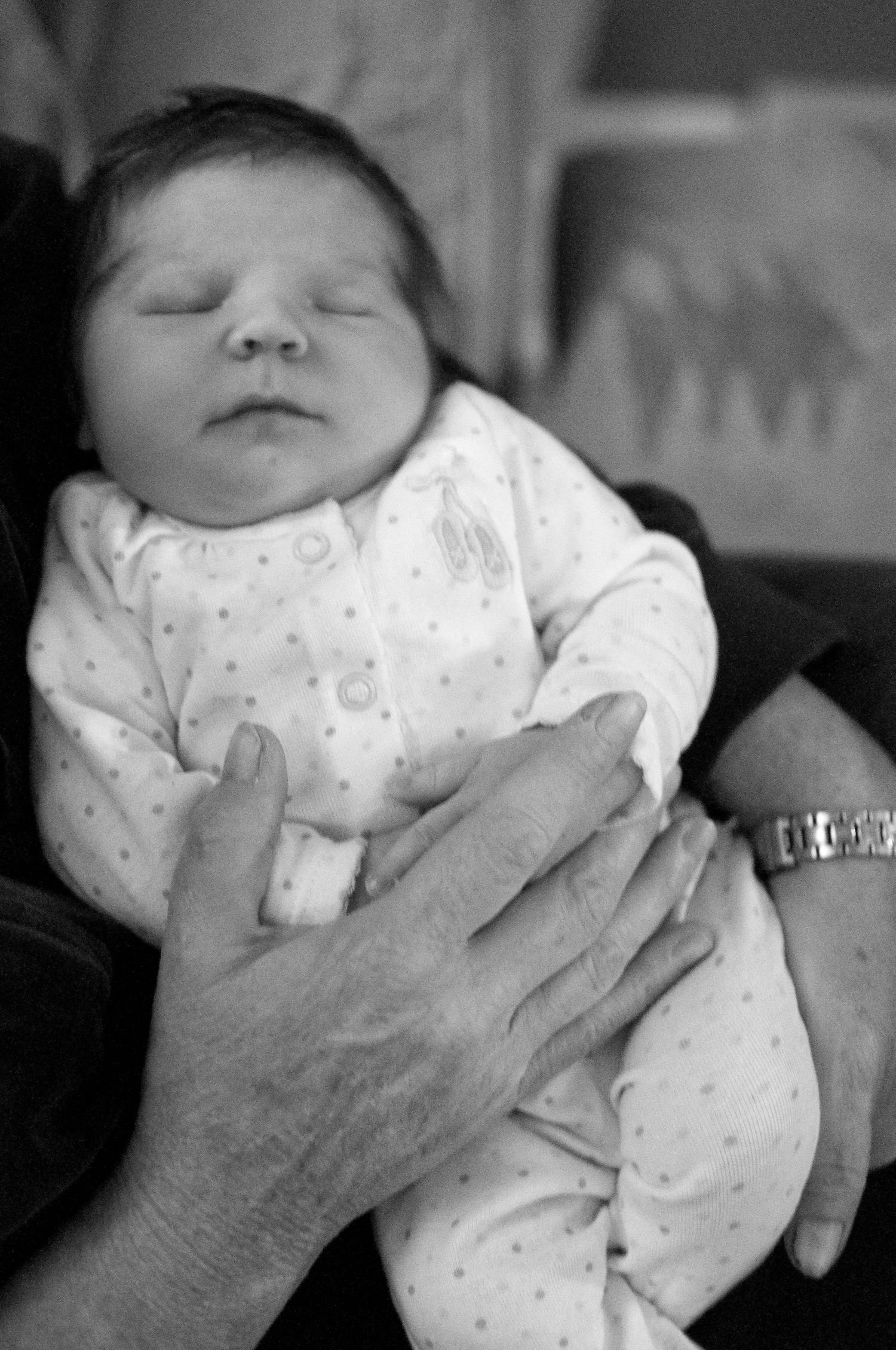 Black and white photo of sleeping newborn in polka dot onesie held by adult hands.