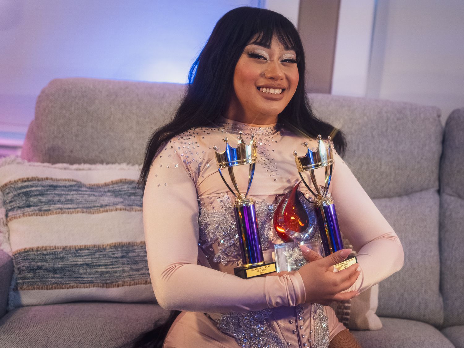 Someone in a white sparkly outfit sits on a couch while holding two blue and gold trophy awards.