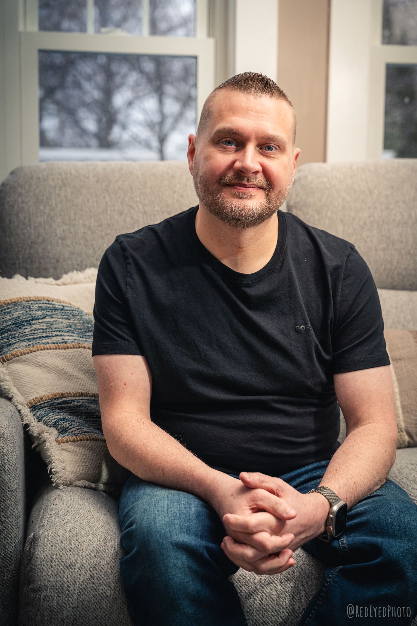A smiling middle-aged man with a beard sits relaxed on a couch, wearing a black t-shirt and jeans.