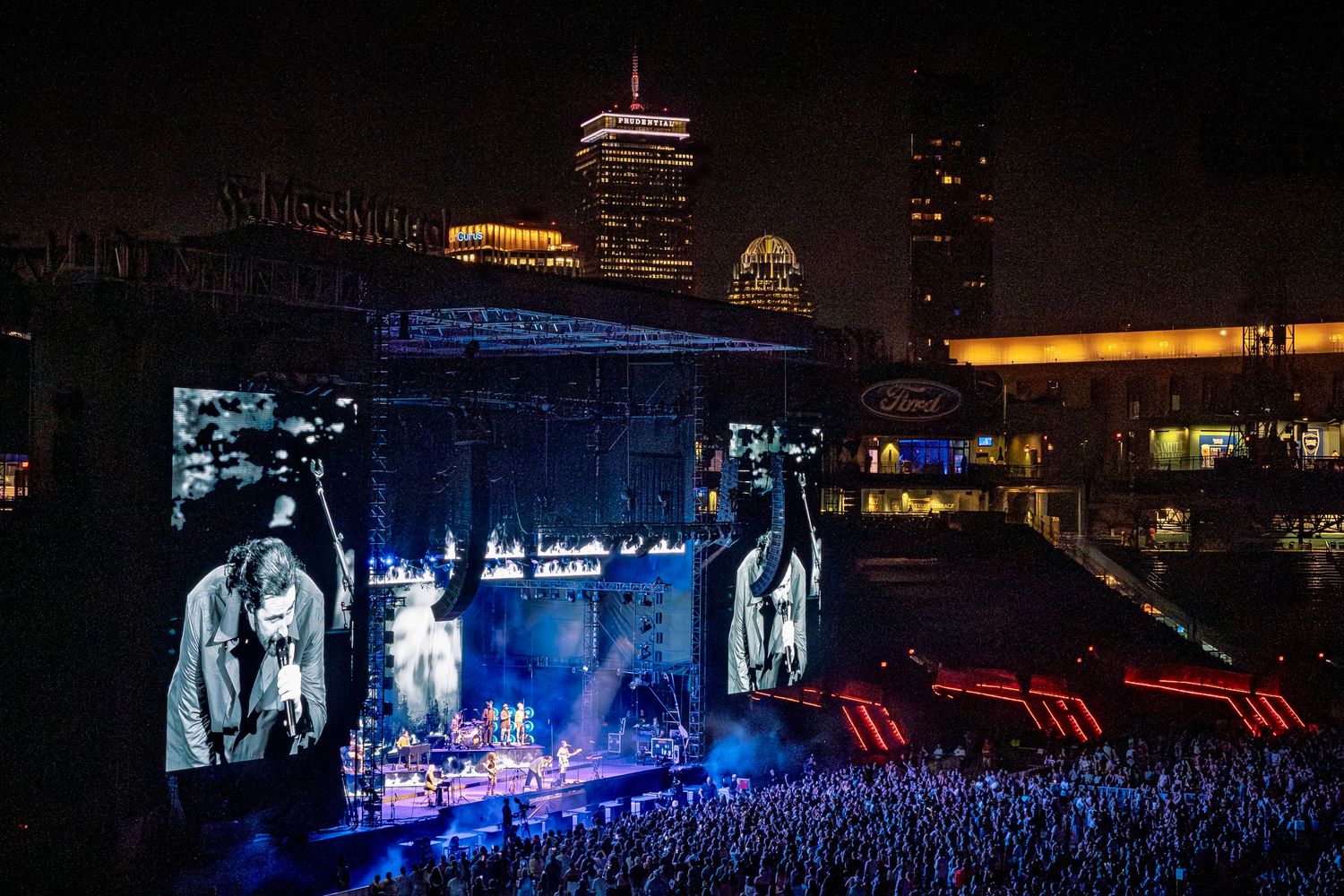 A nighttime concert venue with blue stage lighting illuminating a packed crowd of spectators watching a live performance.