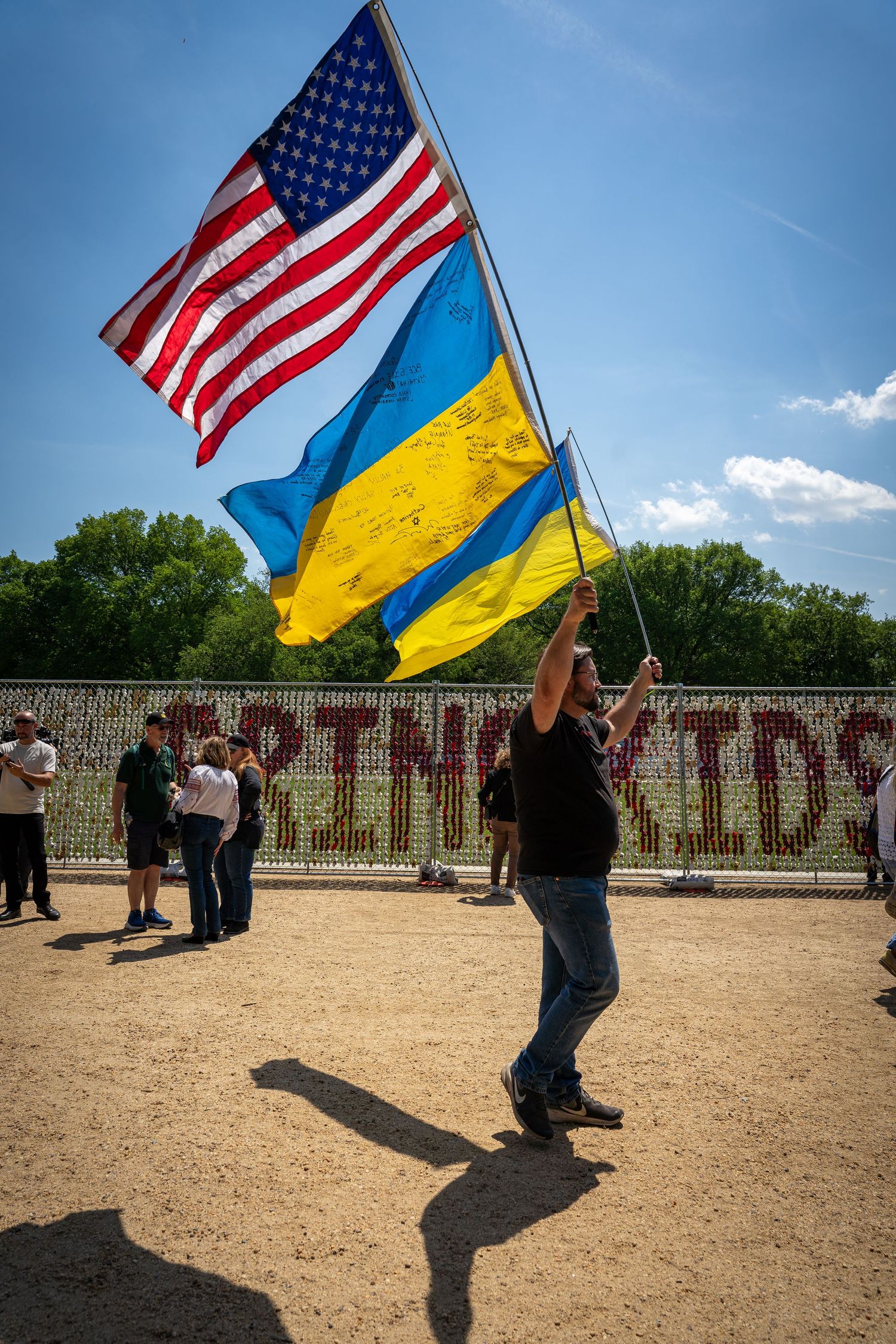 Man waving American and Ukrainian flags outdoors at a sunny rally with a fence in the background covering a section of the National Mall.