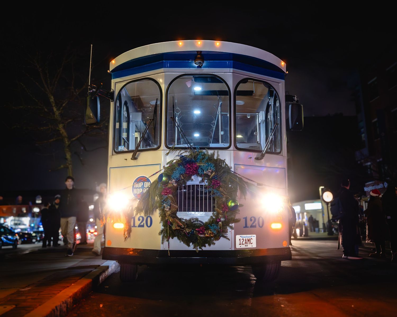 Vintage bus decorated with a Christmas wreath drives down a dark street at night with glowing headlights.