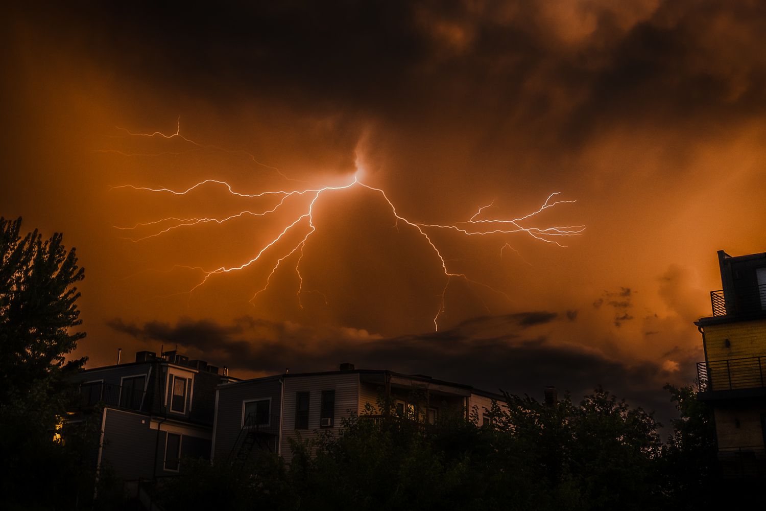 Multiple lightning bolts illuminate a dark stormy sky above silhouetted houses and trees during a dramatic thunderstorm.