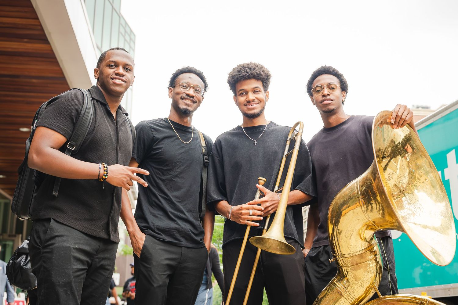 Four young musicians in black attire pose outdoors with a trombone and tuba at a jazz event.