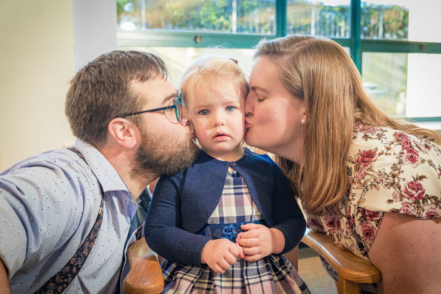Parents kissing their toddler daughter on the cheeks simultaneously while she looks at the camera.