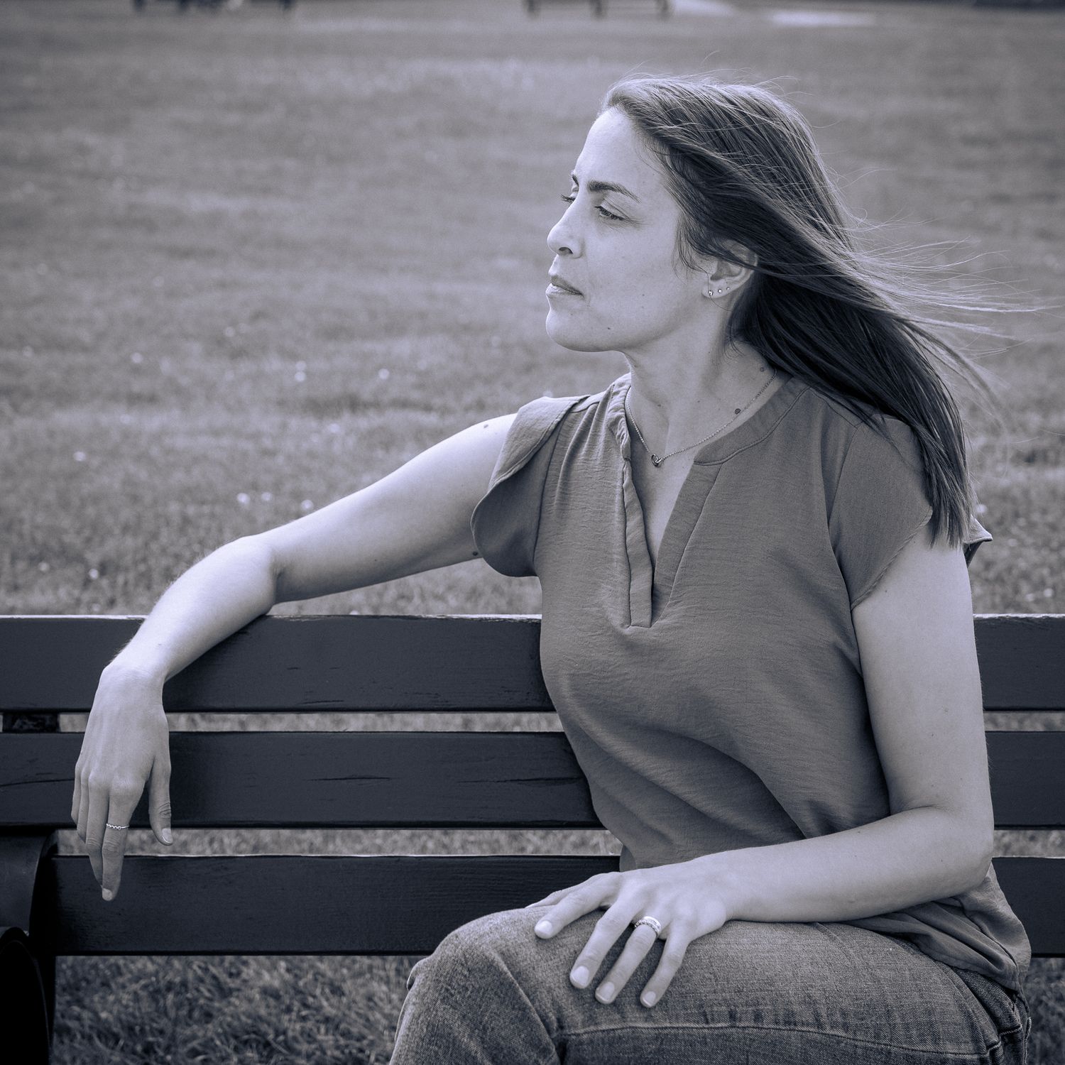 A peaceful contemplative figure sits alone on a park bench with windswept hair in a black and white artistic photo.