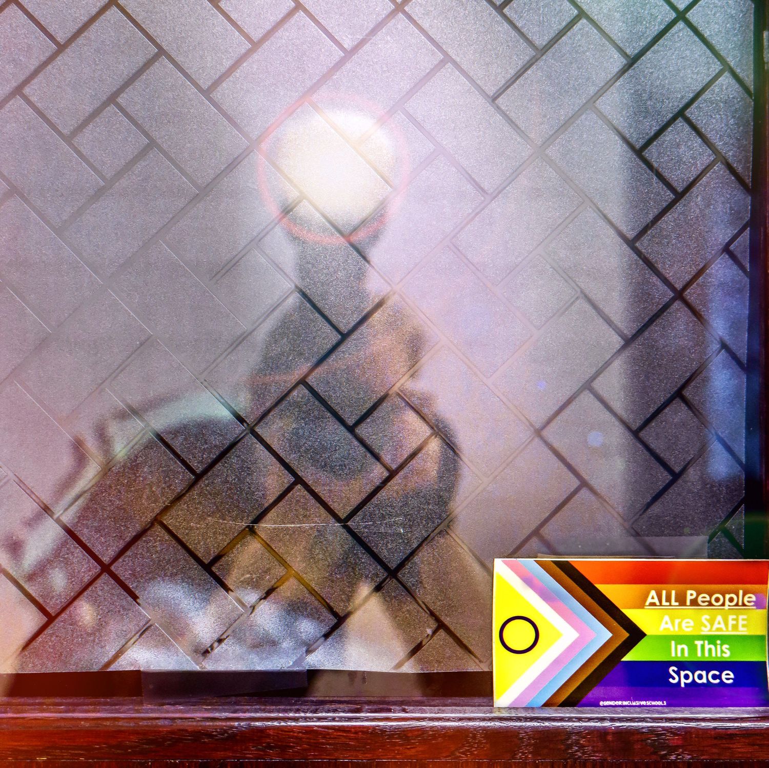 A blurred silhouette behind textured glass with a rainbow pride sticker visible in the corner.