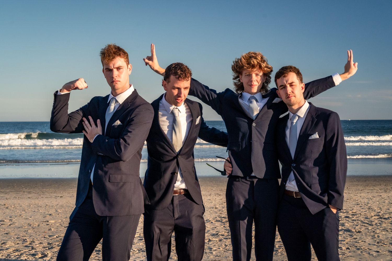 Four people in dark suits strike playful poses together on a sunny beach with ocean waves in the background.