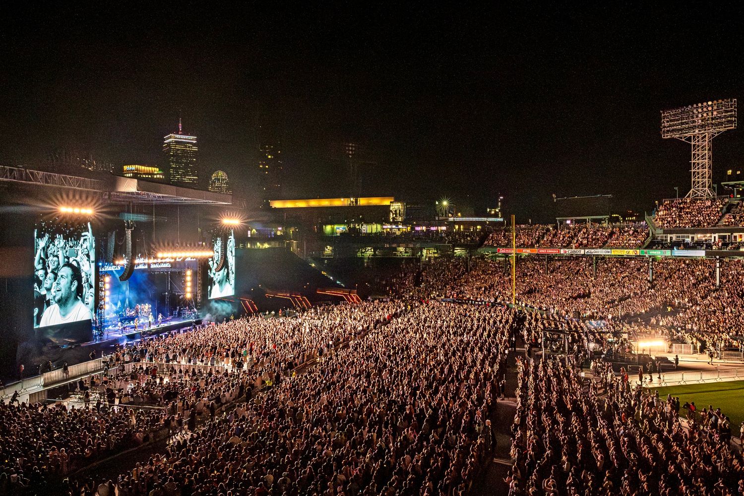 Massive crowd fills outdoor stadium at night for concert with large video screens and city skyline in background.