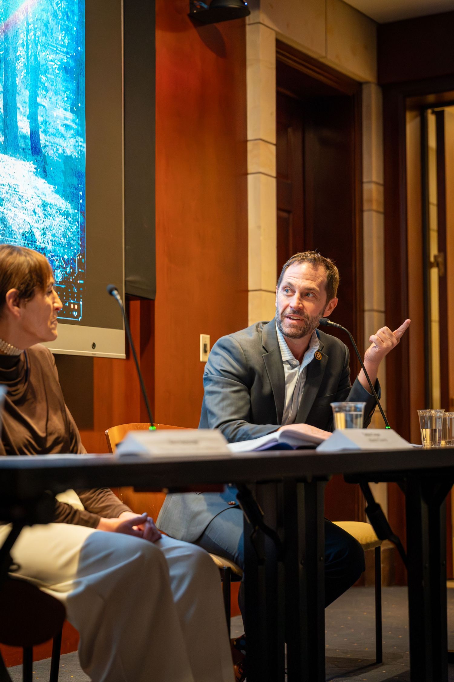 Two professionals engaged in discussion at a panel event, man in suit gesturing while speaking at a conference table.