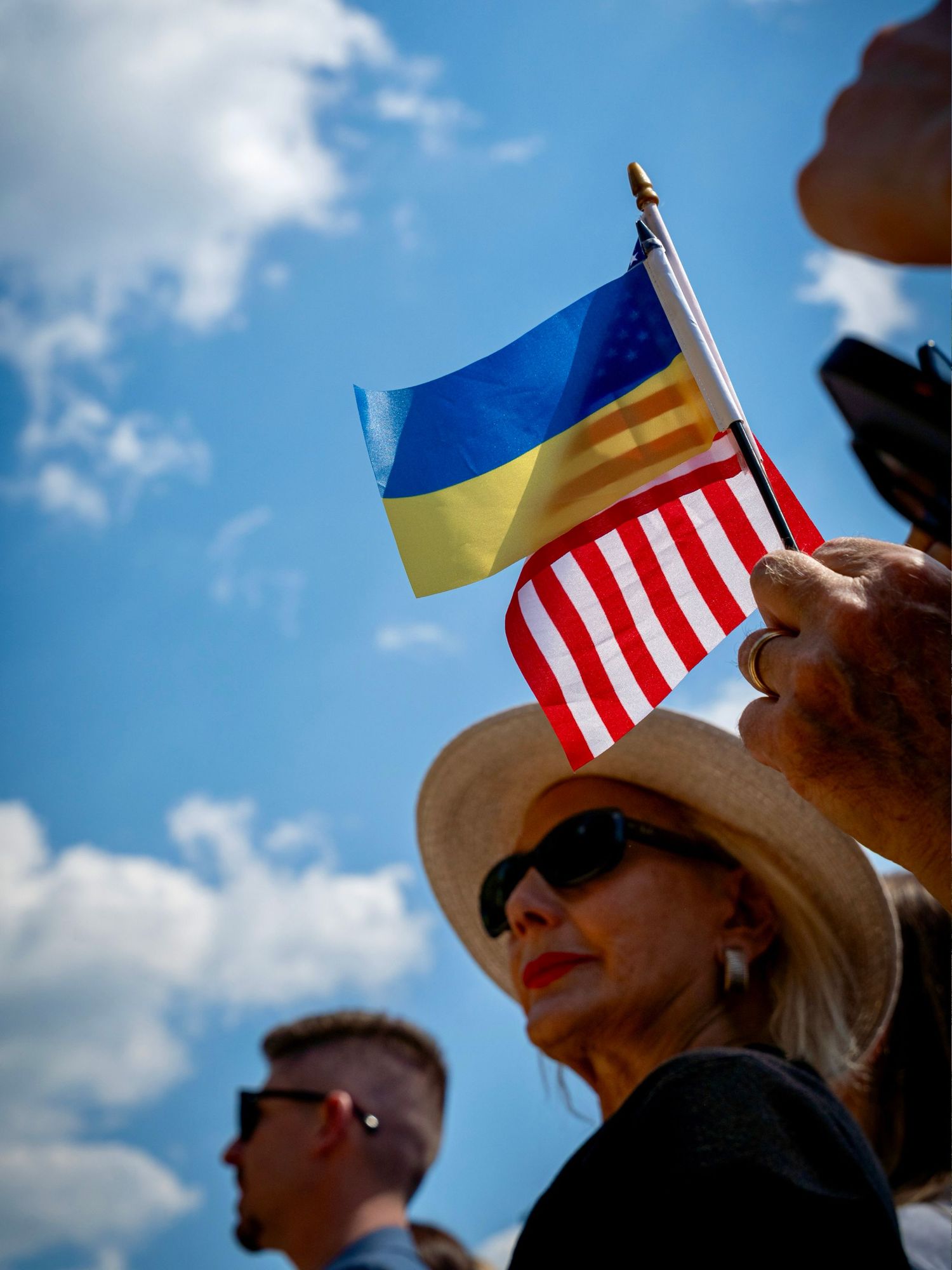 Woman in hat holding Ukrainian and American flags together against a blue sky at a solidarity rally.