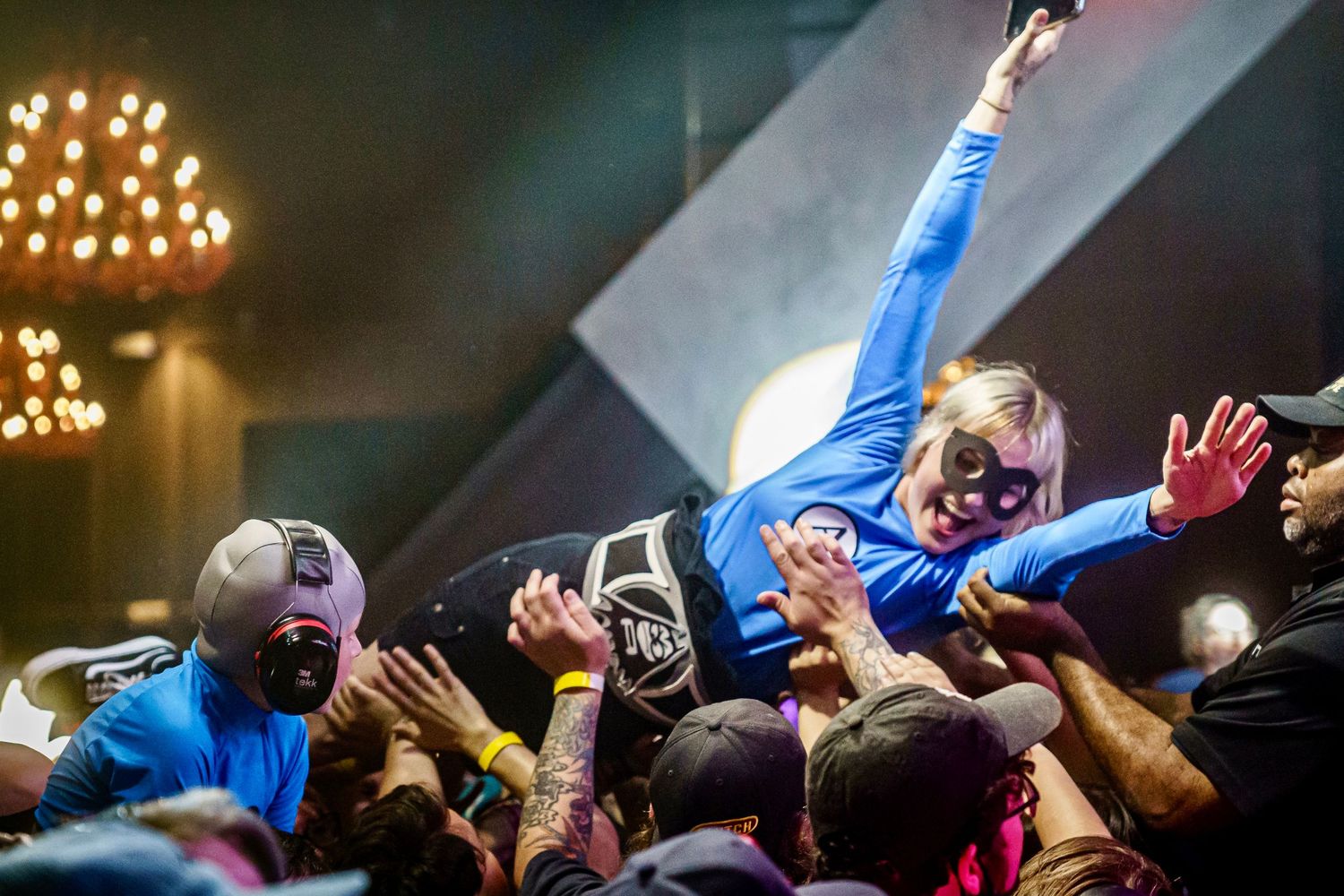 Crowd surfer in blue jumpsuit carried by concert audience in a vibrant, energetic live music venue.