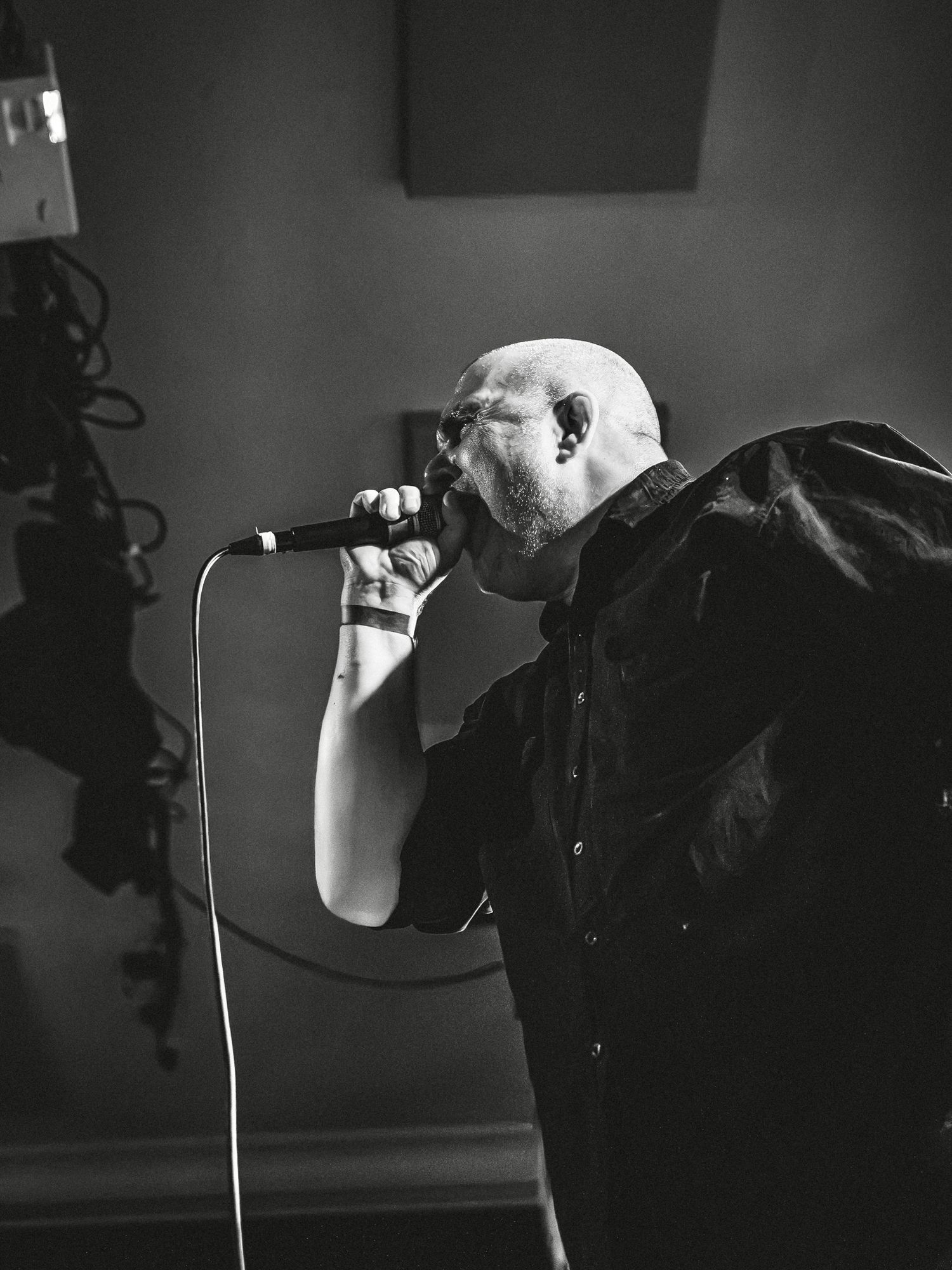 Bald male vocalist performing live, singing intensely into a microphone, wearing black shirt, black and white photo.