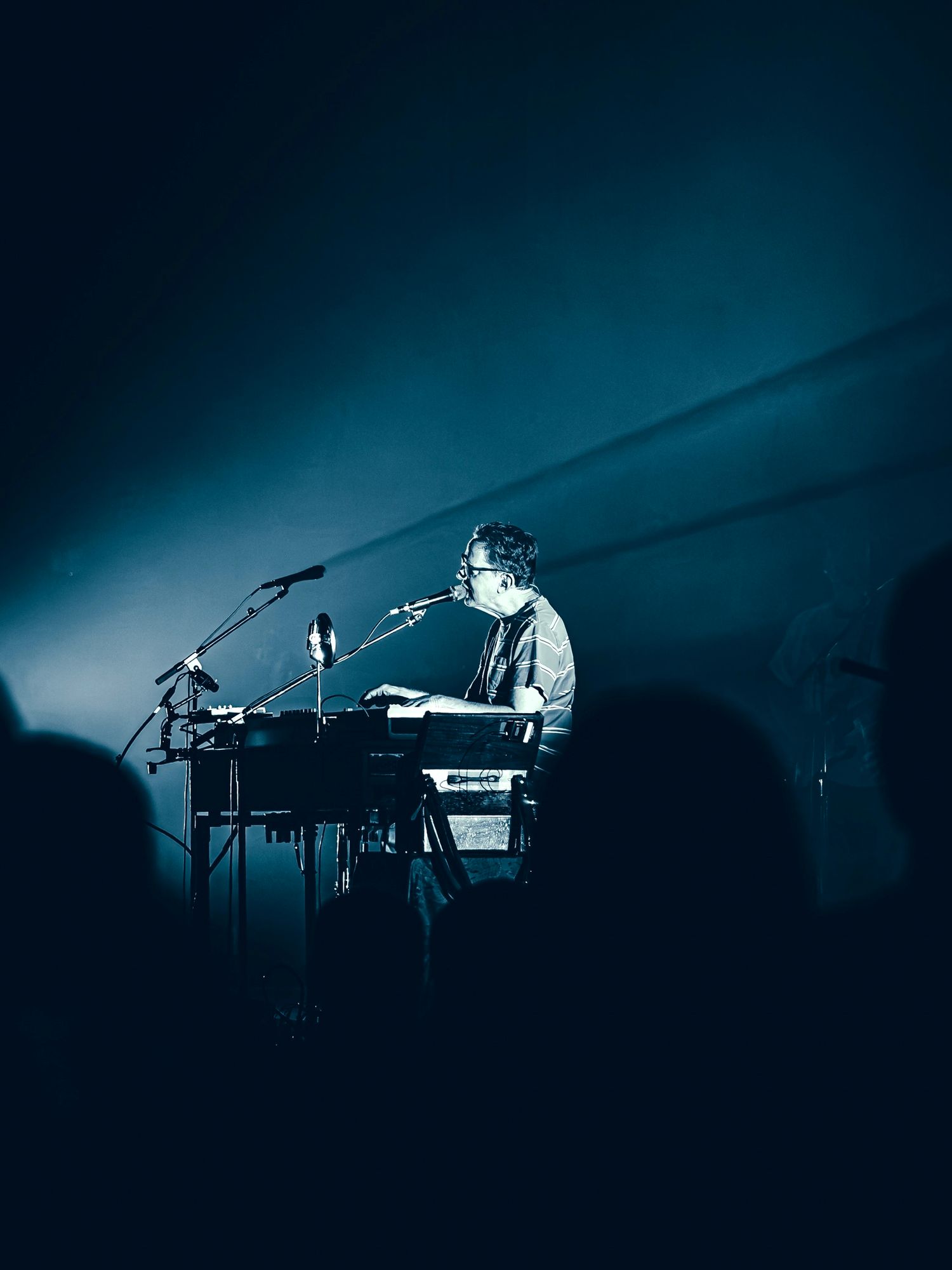 Musician performing on keyboard and synthesizer on dimly lit stage with blue lighting during live concert.
