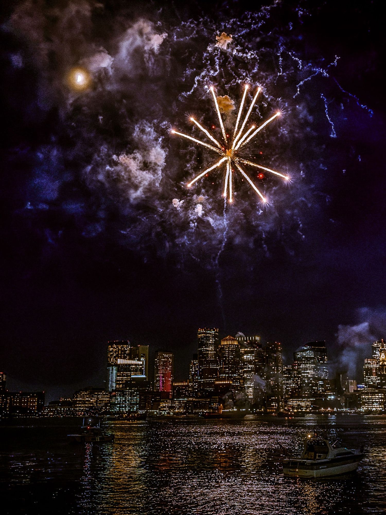 Lightning and fireworks illuminate a city skyline at night with reflections shimmering across dark water.
