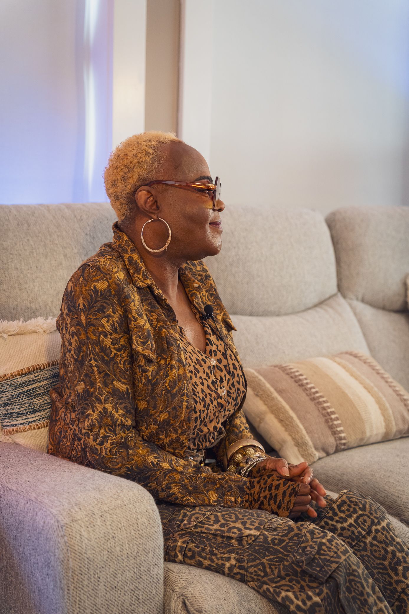 Smiling person wearing glasses and animal print outfit sits comfortably on a gray couch with decorative pillows.