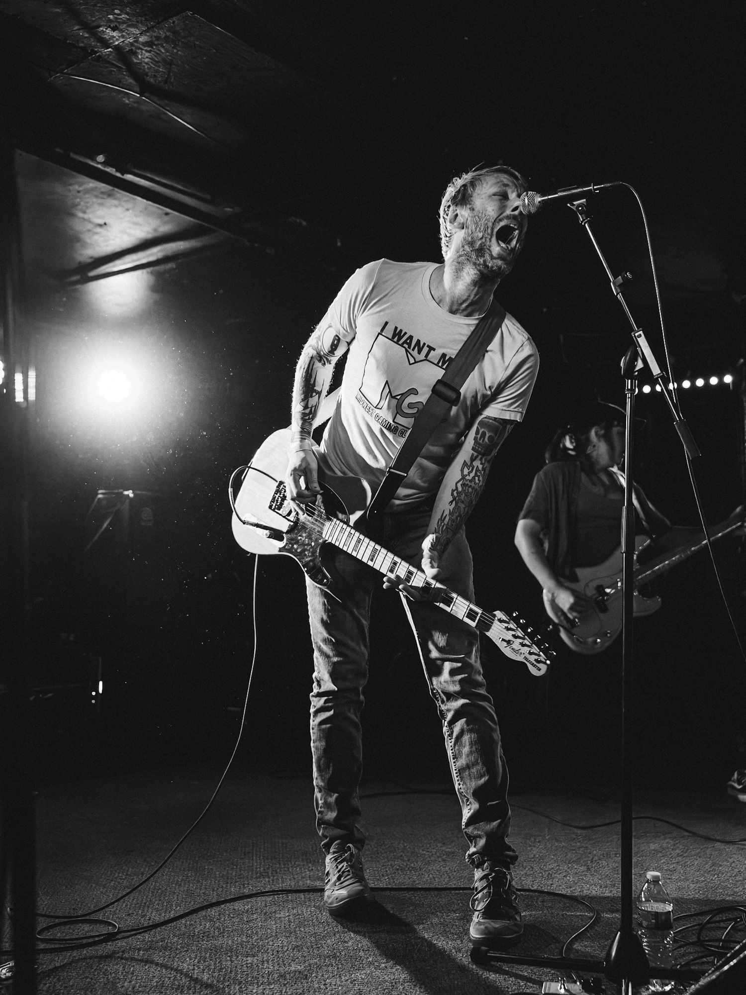 Tattooed guitarist singing into microphone on stage in black and white live show.  (Band: Direct Hit!)