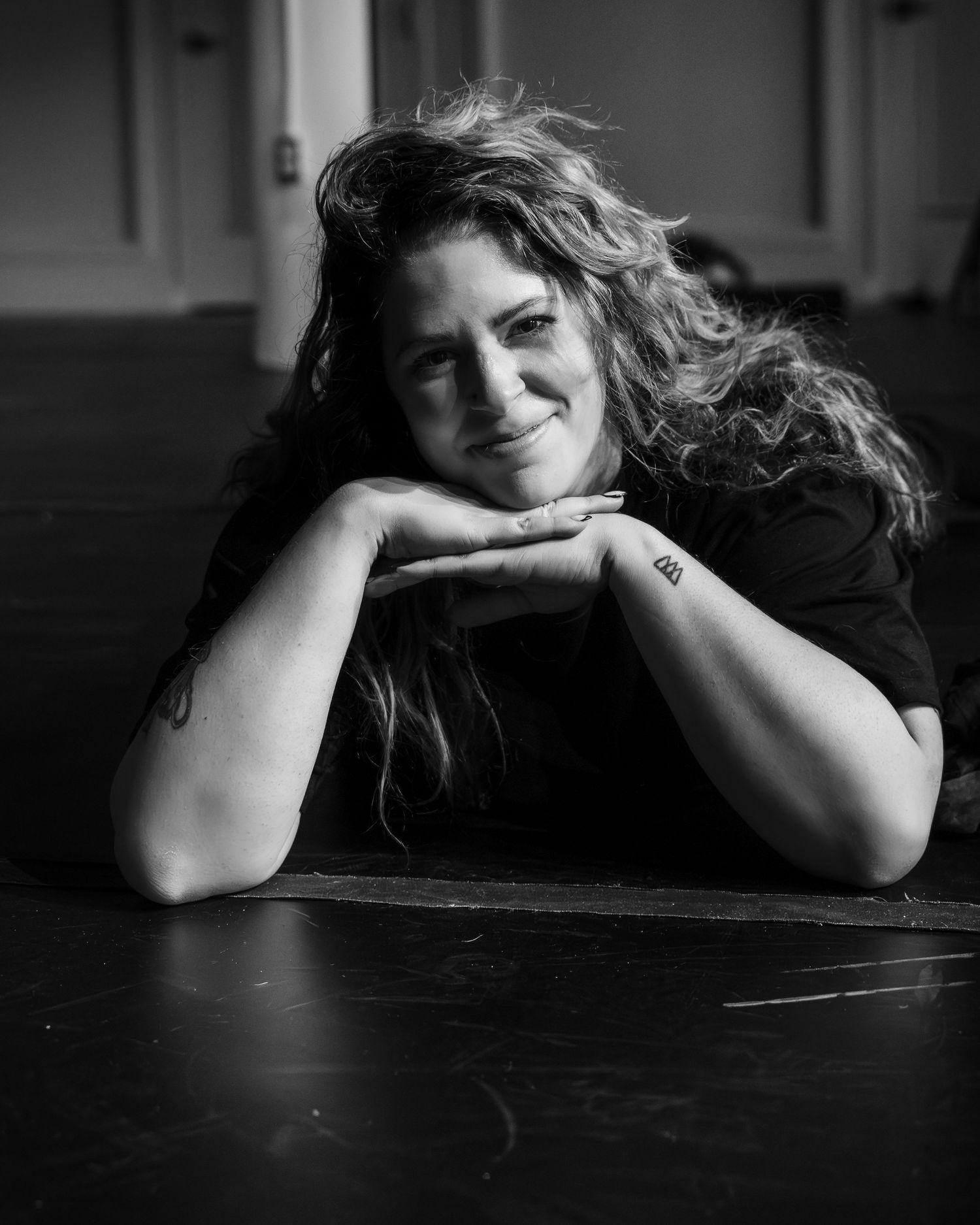 Black and white portrait of a smiling woman with curly hair resting chin on hands, showing a small tattoo.