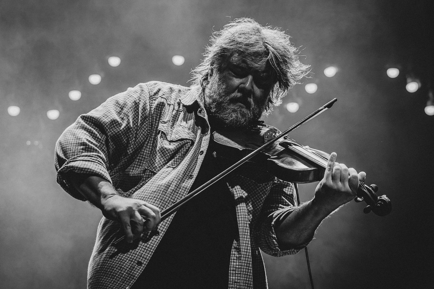 Bearded man playing violin on stage in dramatic black and white concert photography with stage lights.