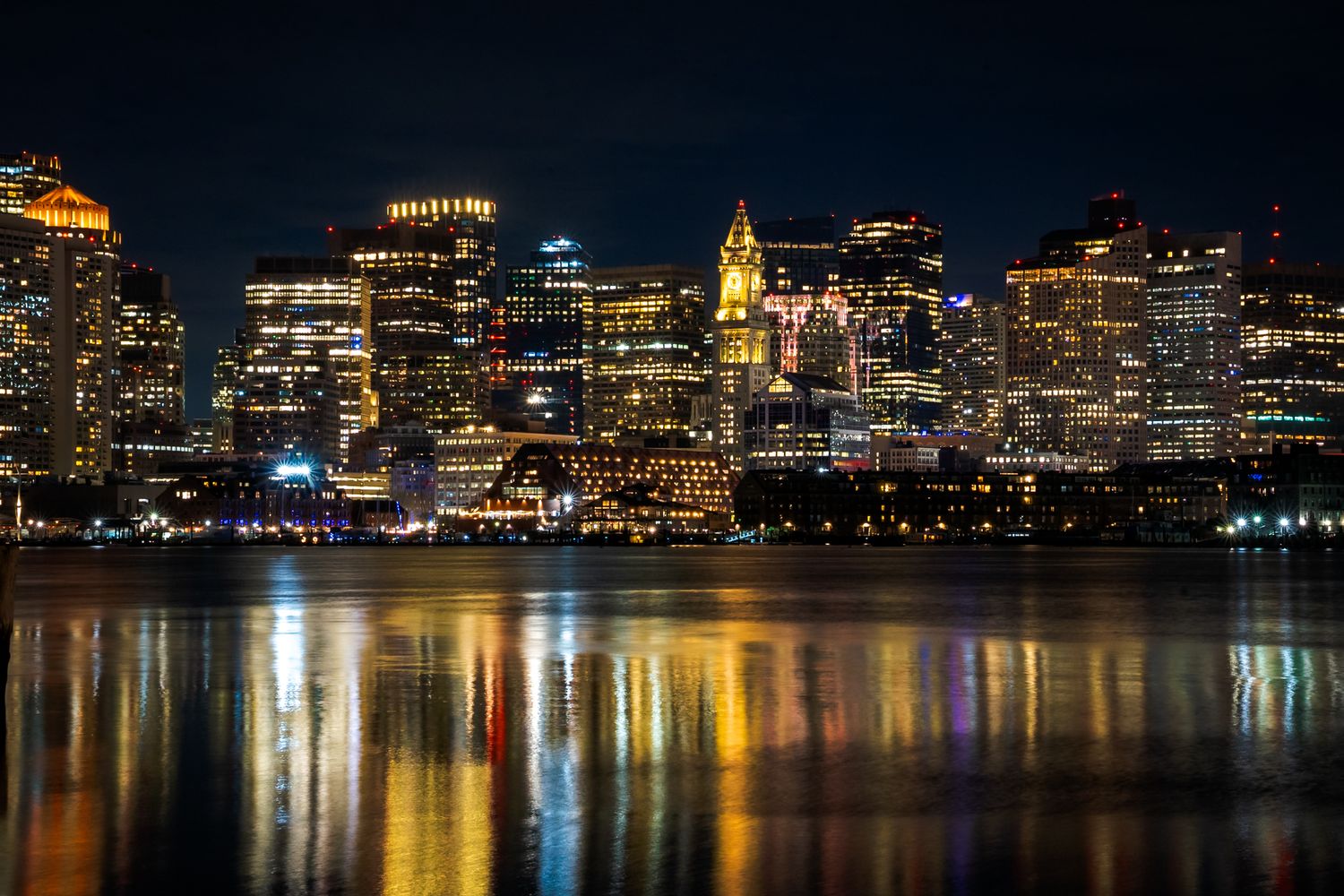 Boston skyline reflects in Charles River at night with glowing city lights illuminating the waterfront.