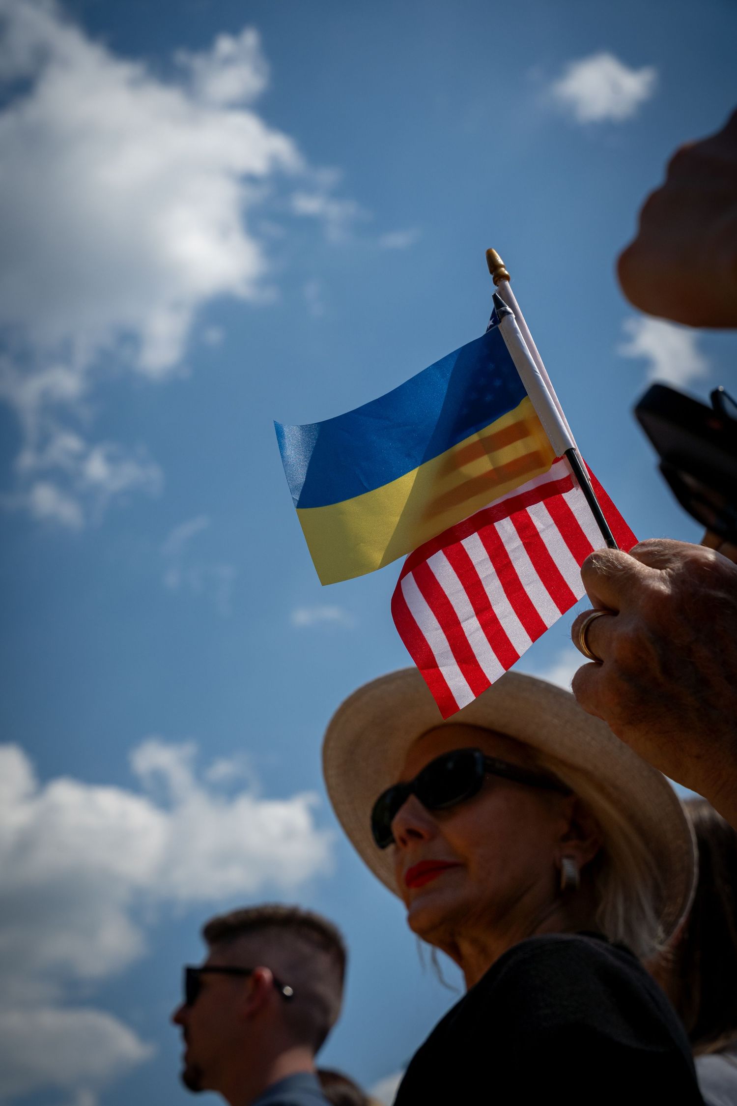 Woman in sun hat holds Ukrainian and American flags at outdoor rally against blue sky in the heart of the National Mall in Washington, D.C.