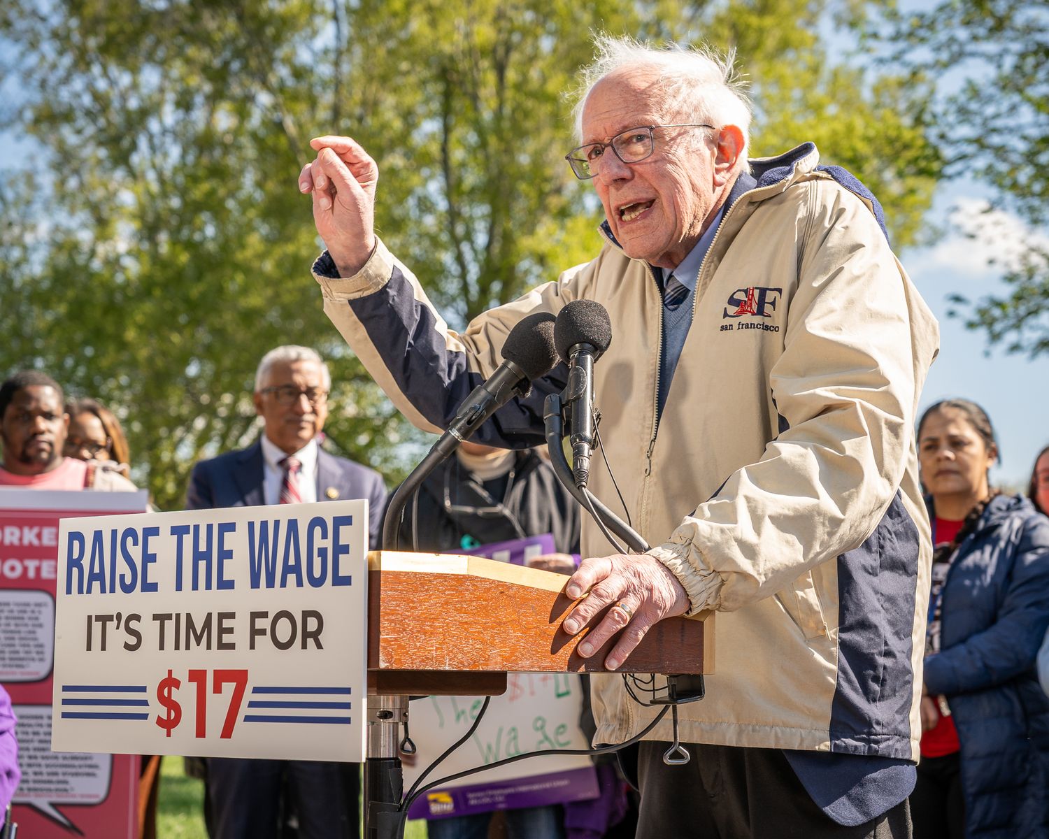 A person speaks passionately behind a podium at a rally with 'Raise the Wage' protest signs visible in the background.