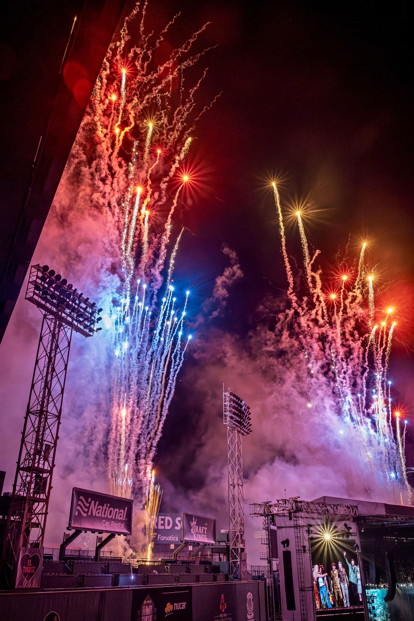 Vibrant fireworks illuminate a stadium at night with colorful bursts of red, blue and yellow lights against a dramatic cloudy sky.