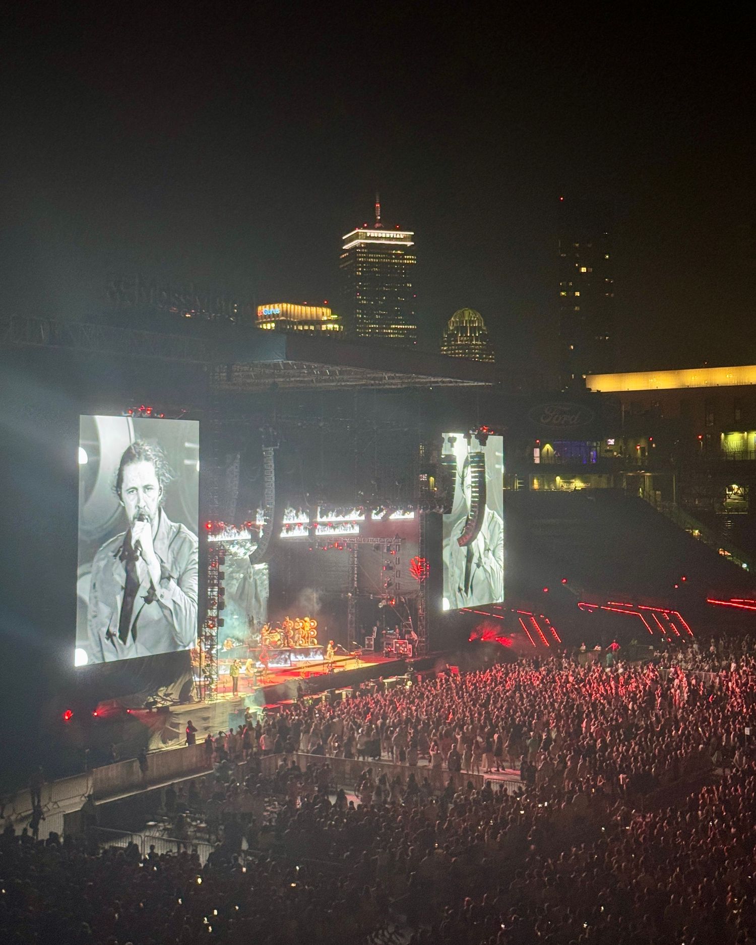 Large concert venue with video screens showing a performer and a massive crowd beneath the Boston skyline at night.